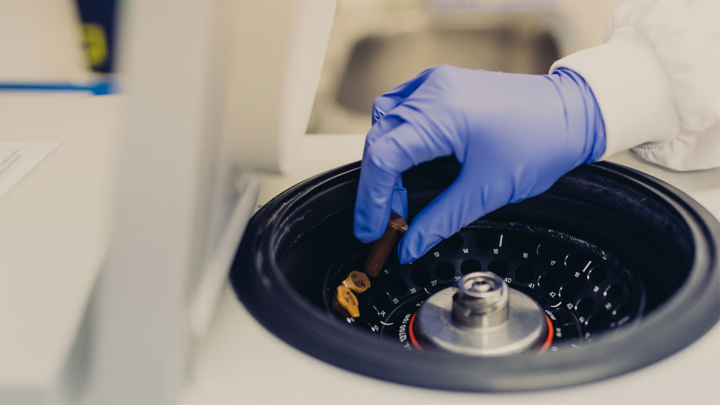 The image showcases a close-up view of a scientist's hand, gloved in blue, carefully placing or retrieving a vial into a centrifuge. The centrifuge appears to be a high-precision laboratory equipment, used for separating substances based on density by spinning them at high speed. The scientist is wearing a white lab coat, indicating a professional laboratory setting. This scene captures a moment of meticulous laboratory work, emphasizing the expertise and precision in bioanalysis. It highglights the company's focus on high-quality, precise analytical services in the field of bioanalysis.