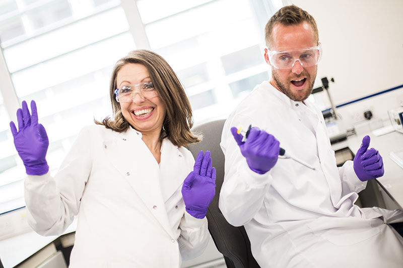 female and male scientist weating white coats, glasses and purple gloves smiling and doing a thumbs up
